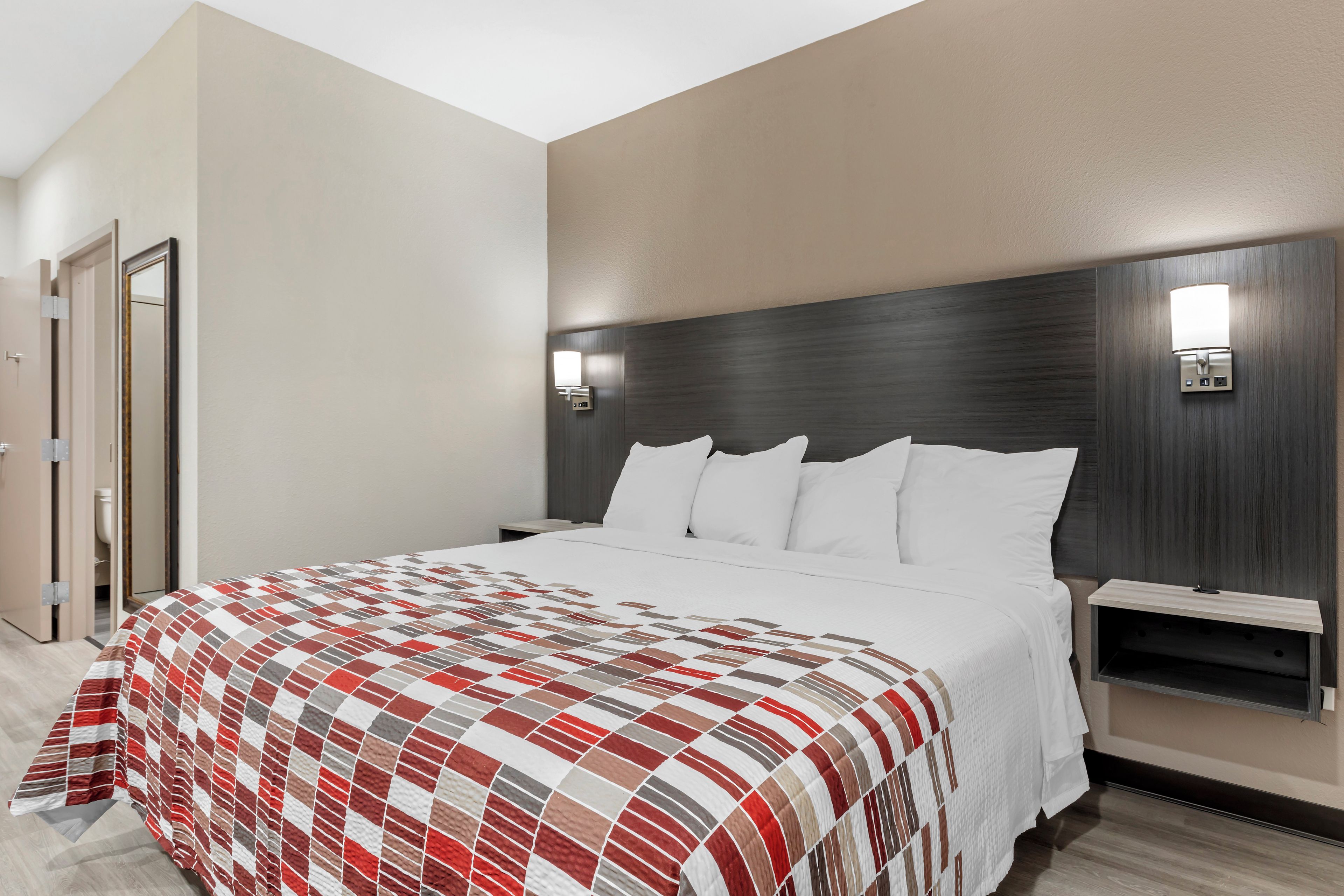 A hotel room with a large bed, a headboard with integrated lighting, a bedside table, and a door leading to a bathroom.