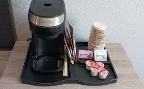 A room with a black coffee maker, a tray with sugar packets, a stack of paper cups, and a container of coffee beans.