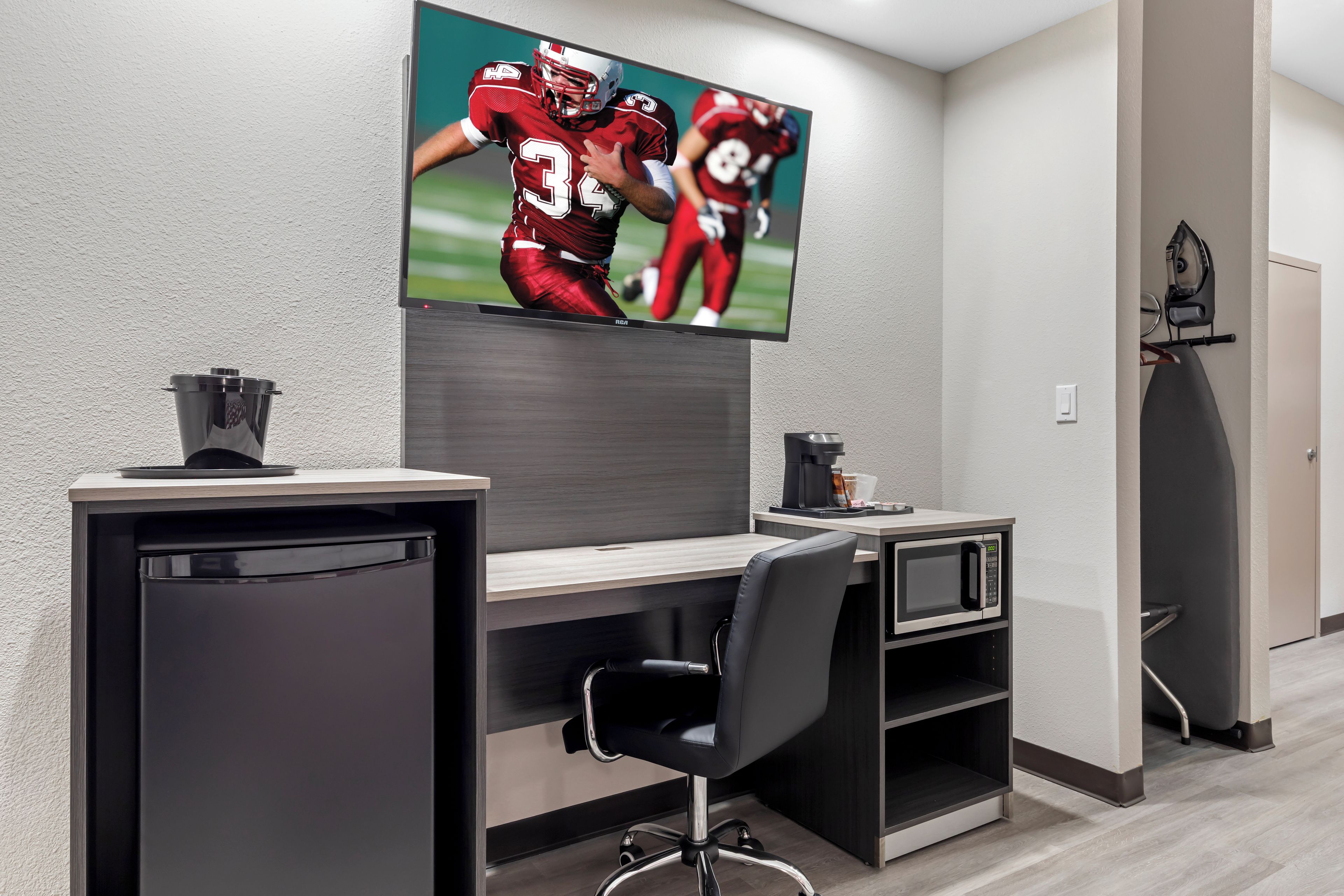 Un espacio de oficina moderno con una televisión de pantalla plana grande montada en la pared, un escritorio con una silla, una cafetera y un cubo de basura.