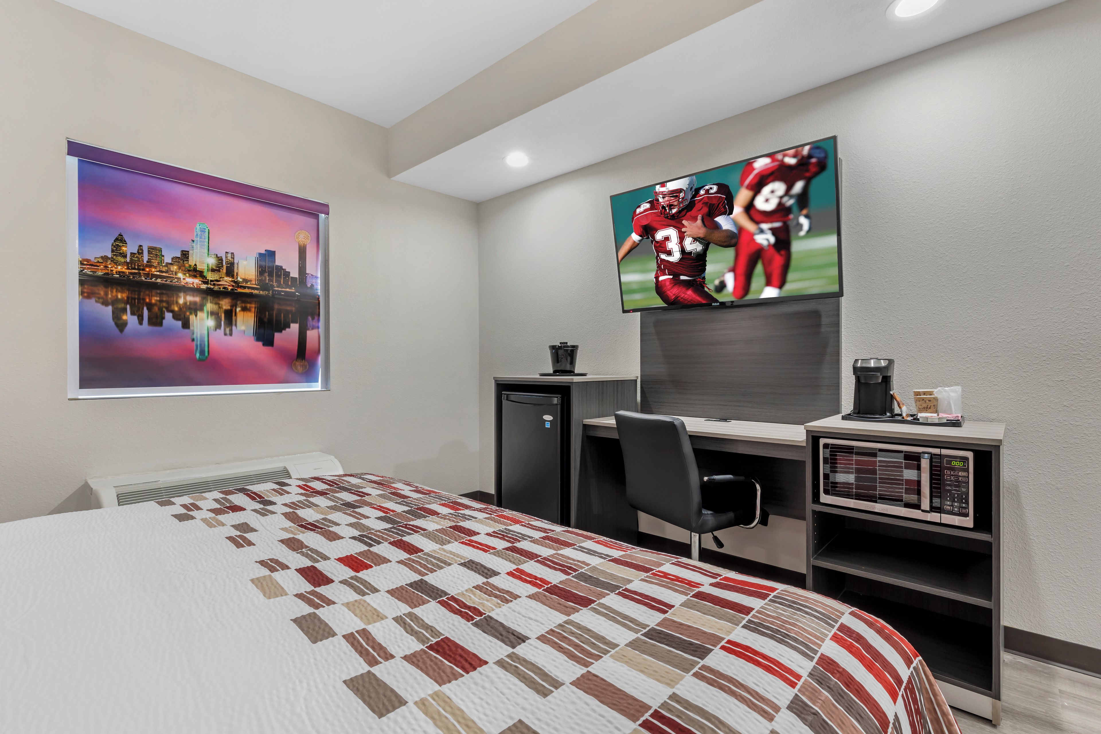 A hotel room with a bed, a desk with a computer, a TV mounted on the wall, and a window with a cityscape view.