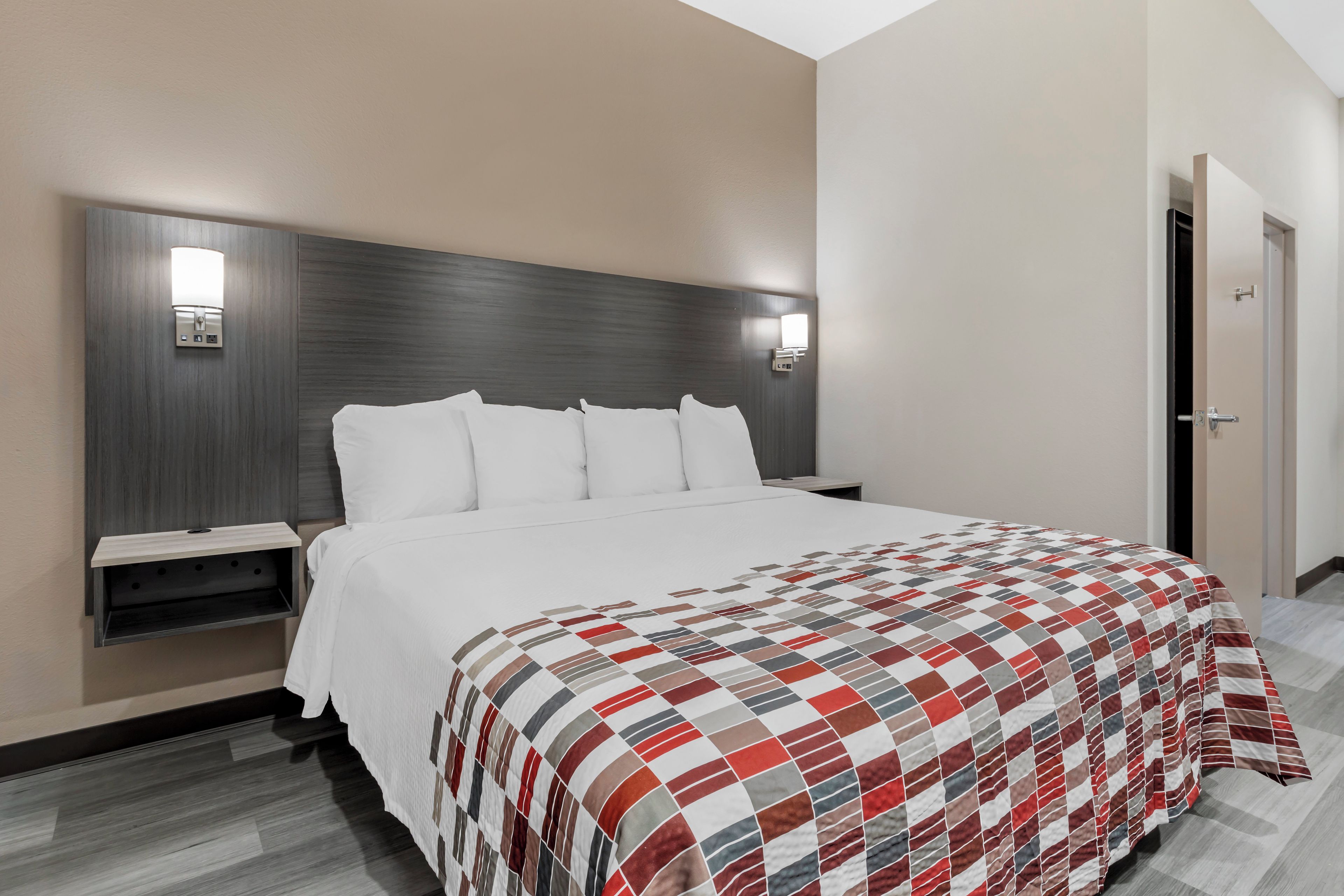 Una habitación de hotel con una cama grande, una mesita de noche, una lámpara de pared y una puerta.