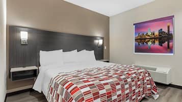 A hotel room with a bed, a nightstand, a wall-mounted air conditioner, and a cityscape painting.