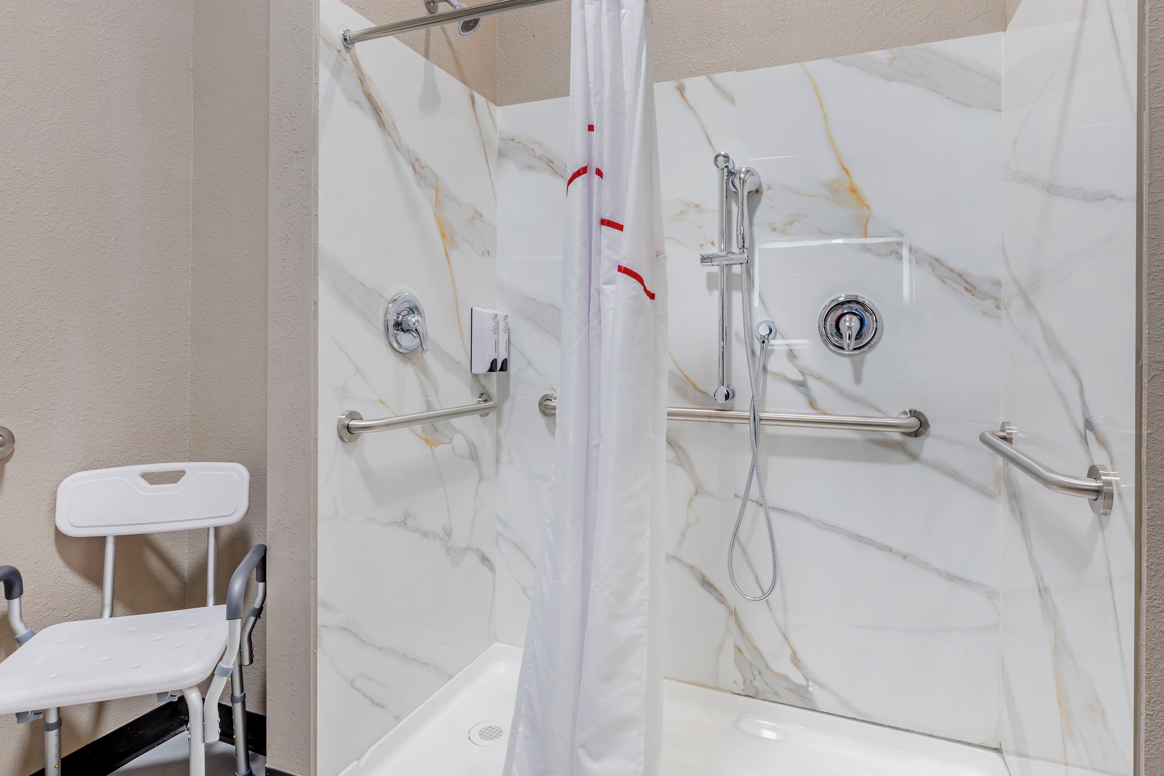Un baño con ducha, barras de apoyo y una silla blanca.