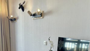 Huoneessa on valkoinen TV-taso, jossa on televisio, kaukosäädin, ruukkuorhidea ja seinälle kiinnitetyt seinävalaisimet.