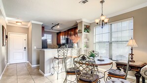 Una cocina amplia con una isla central, un comedor con una mesa redonda y sillas metálicas, y una zona de estar acogedora con un otoman tapizado.