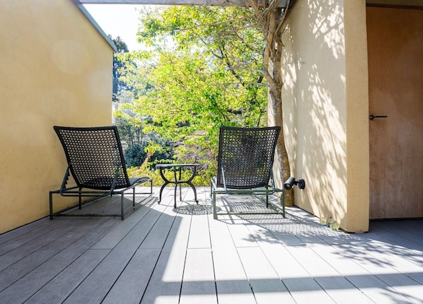 A covered outdoor area with two chairs and a small table, a door, and a view of trees and greenery.
