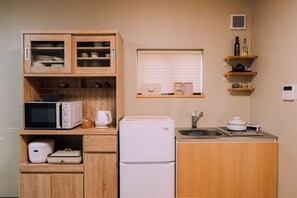 A compact kitchen with wooden cabinets, a microwave, a rice cooker, a refrigerator, a sink, and a window with blinds.