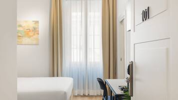Une chambre d’hôtel de taille réduite, comprenant un lit, un bureau, une chaise, une plante en pot et une fenêtre avec des rideaux.