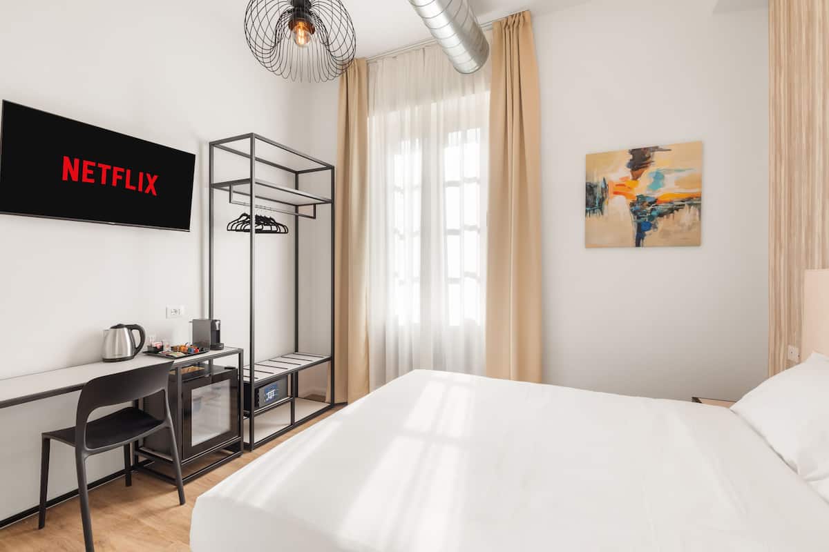 A modern hotel room with a large bed, a desk with a chair, a TV displaying 'NETFLIX', a painting on the wall, and a window with curtains.