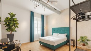 A modern hotel room with a teal bed, a potted plant, a desk with a laptop, and a hanging light fixture. - Adelante Rooms (Rome)