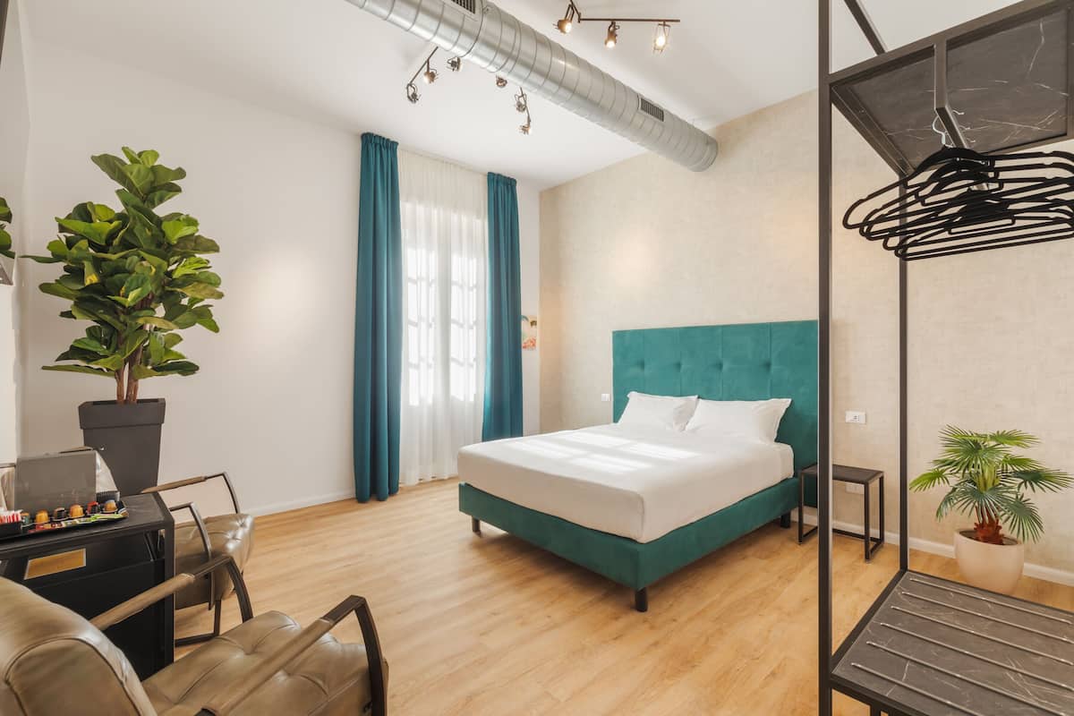 A modern hotel room with a teal bed, a potted plant, a desk with a laptop, and a hanging light fixture.