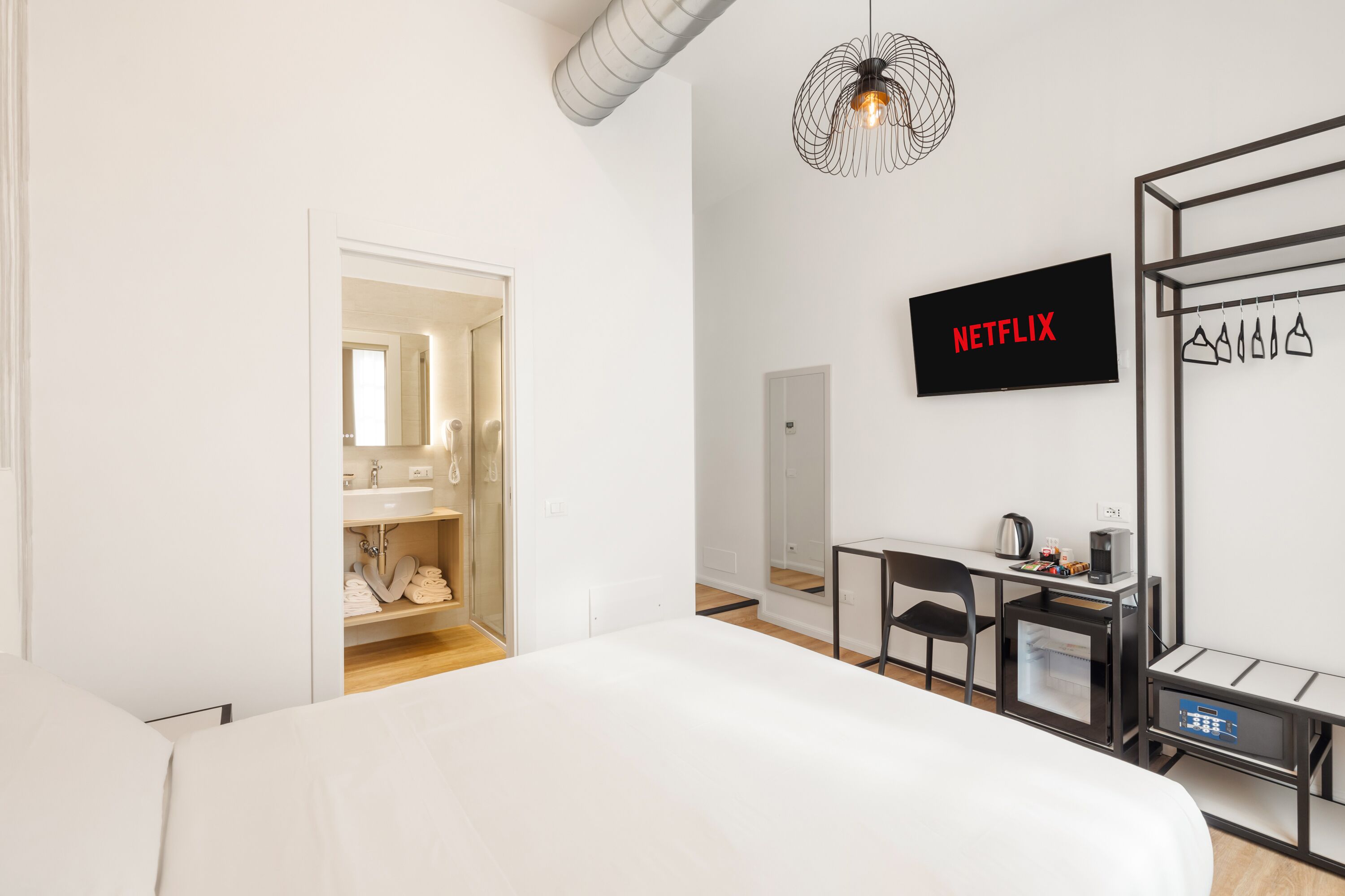 Een moderne hotelkamer met een groot bed, een bureau met een computer, een televisie waarop 'NETFLIX' staat, en een kledingkast met hangende kleding.