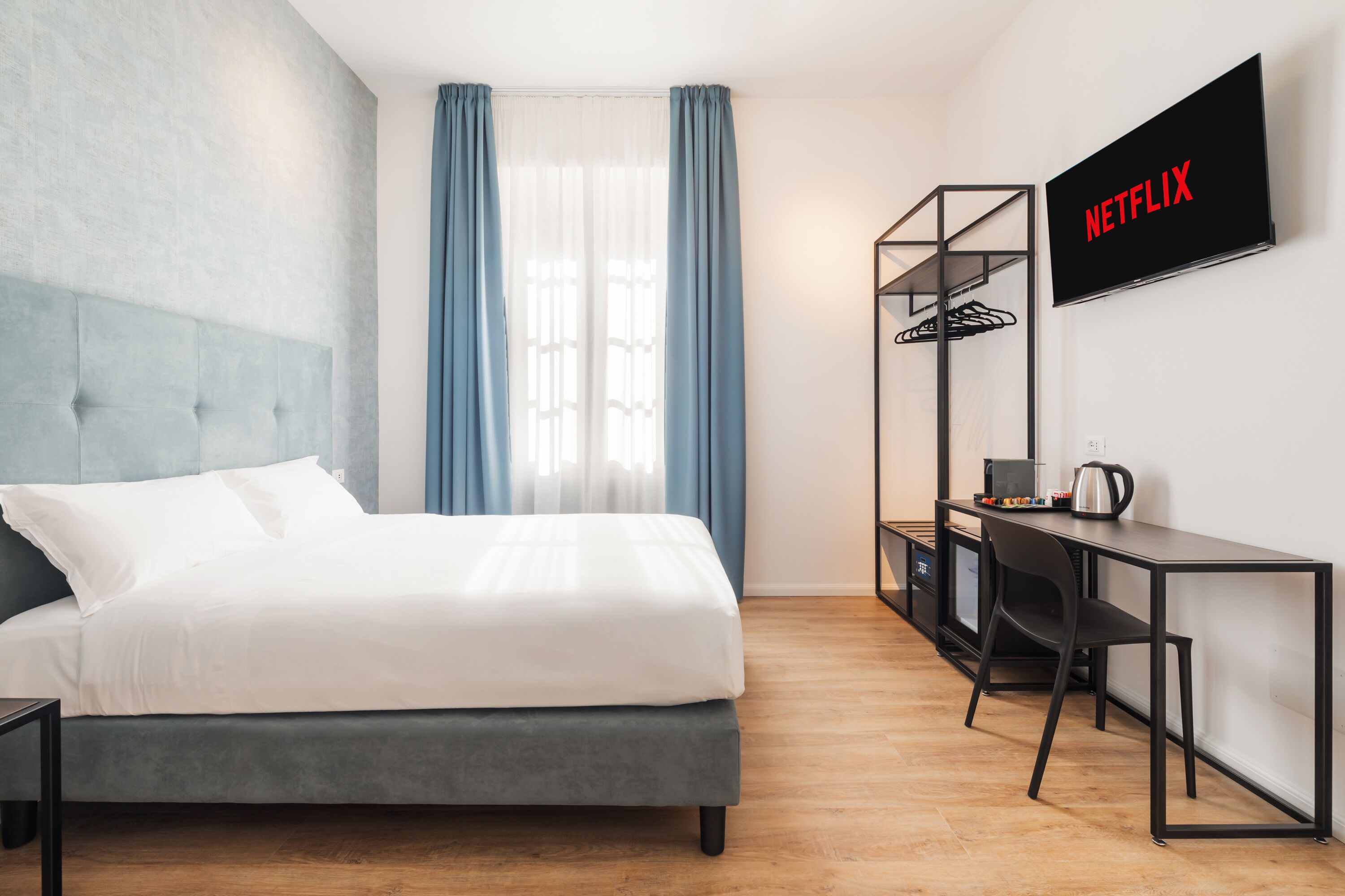 A modern hotel room with a bed, a desk, a chair, a TV, and a wardrobe.
