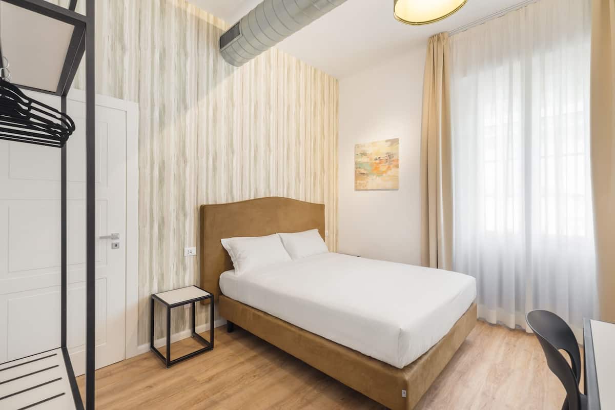 A bedroom with a wooden bed, a small bedside table, a wall-mounted shelf, and a window with sheer curtains.