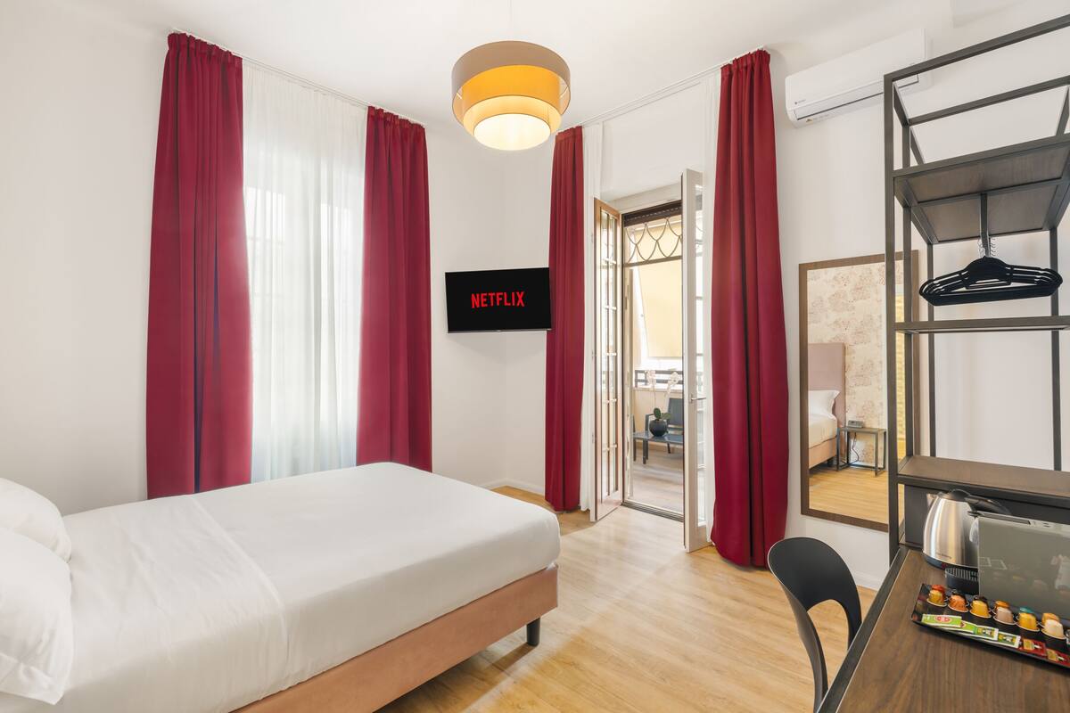 A hotel room with a bed, a desk with a laptop, a TV displaying 'NETFLIX', and a view of the outside through a door.