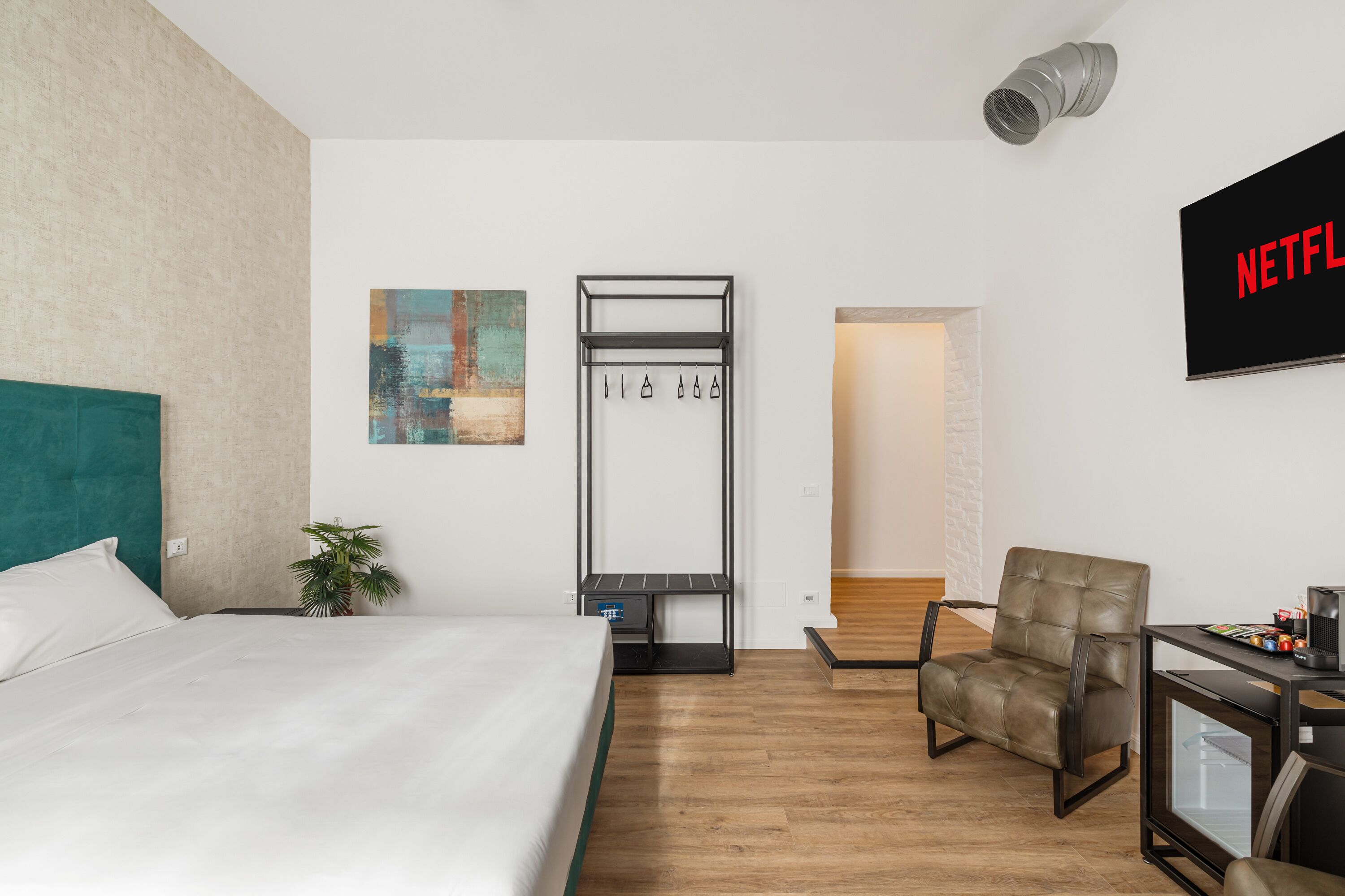 A hotel room with a bed, a chair, a small table, a coat rack, and a wall-mounted TV displaying the Netflix logo.