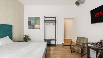 A hotel room with a bed, a chair, a small table, a coat rack, and a wall-mounted TV displaying the Netflix logo.