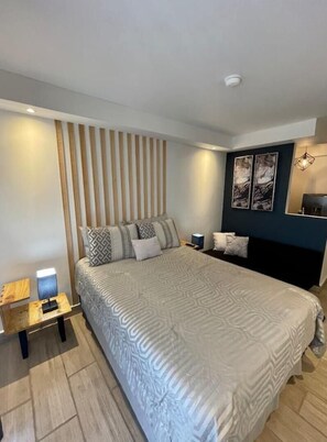 Une chambre moderne avec un grand lit, un mur en lambris de bois, un mur d’accent de couleur foncée agrémenté d’œuvres d’art, et une table de chevet avec une lampe.