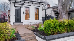 A white house with a black door, two windows with shutters, a small porch with a bench, and a tree in the front yard.