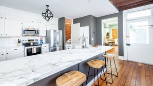 A modern kitchen with white cabinets, a large marble island, wooden stools, and a brick accent wall.