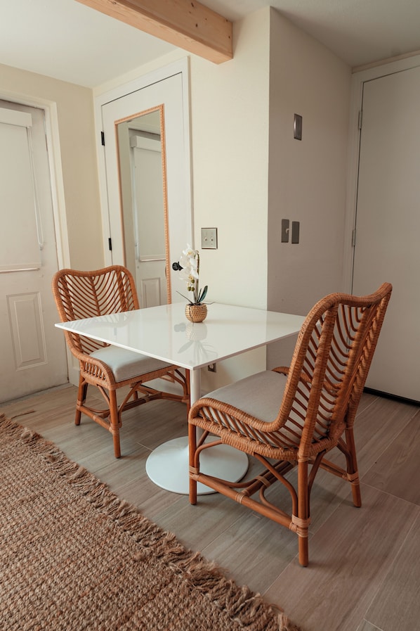 Un comedor con una mesa blanca, sillas de mimbre, una planta pequeña y un espejo en la pared.