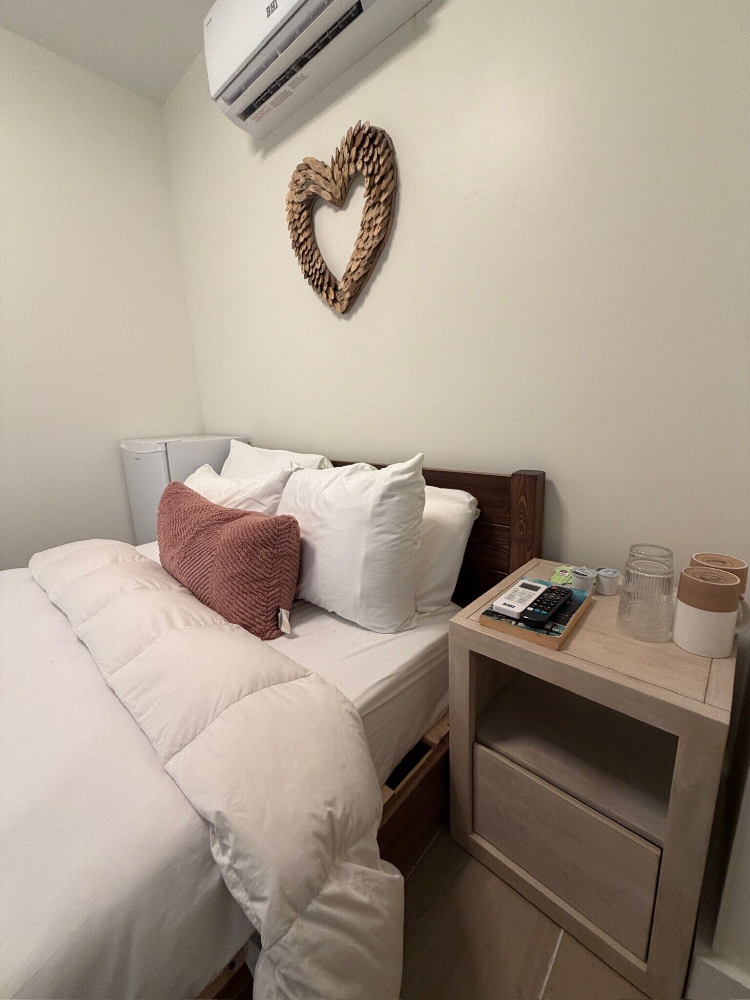 Un lit bien fait, avec une literie blanche et un plaid marron, une table de chevet avec une calculatrice et des lunettes, et une décoration murale en forme de cœur.