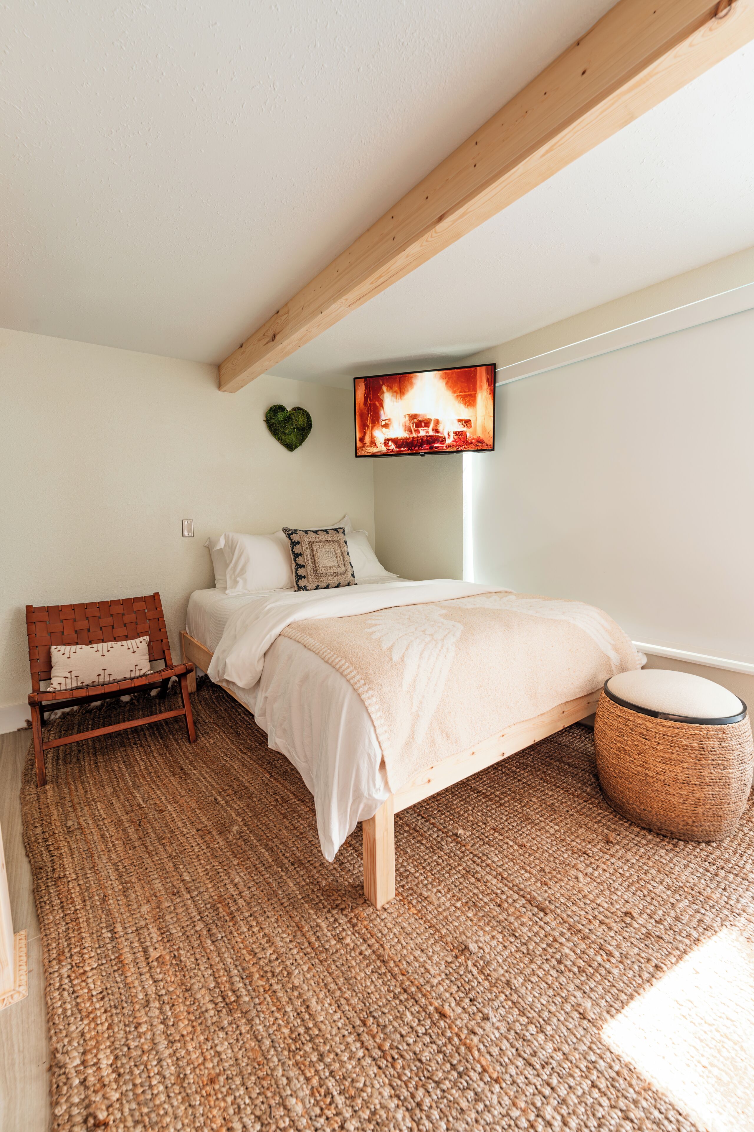 Une chambre à coucher comprenant un lit, une chaise en bois, un pouf en osier, un téléviseur fixé au mur et un objet décoratif en forme de cœur.