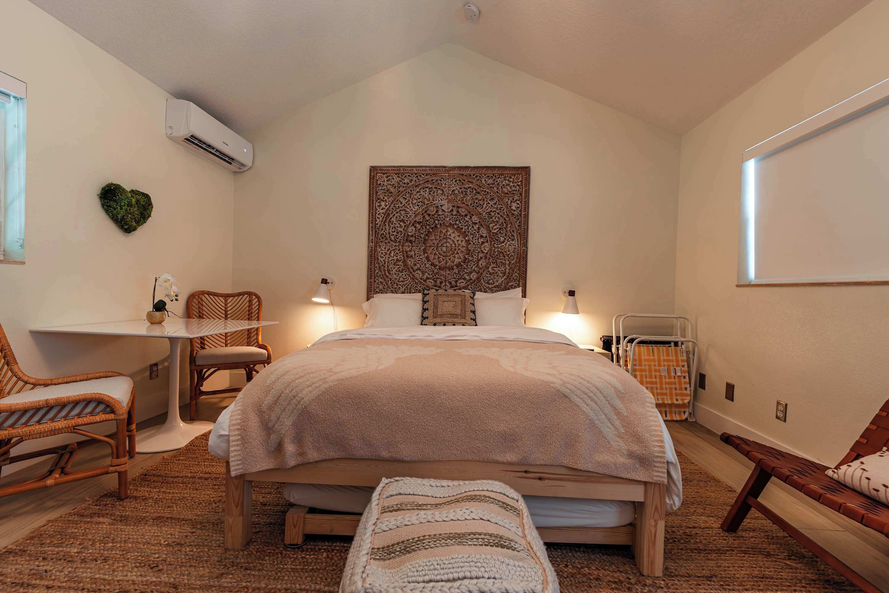 A bedroom with a large bed, a small table, two chairs, a bench, a wall-mounted air conditioner, and a decorative tapestry.
