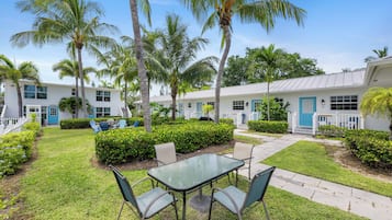 A hotel courtyard with a glass table and chairs, surrounded by palm trees and a well-maintained lawn.