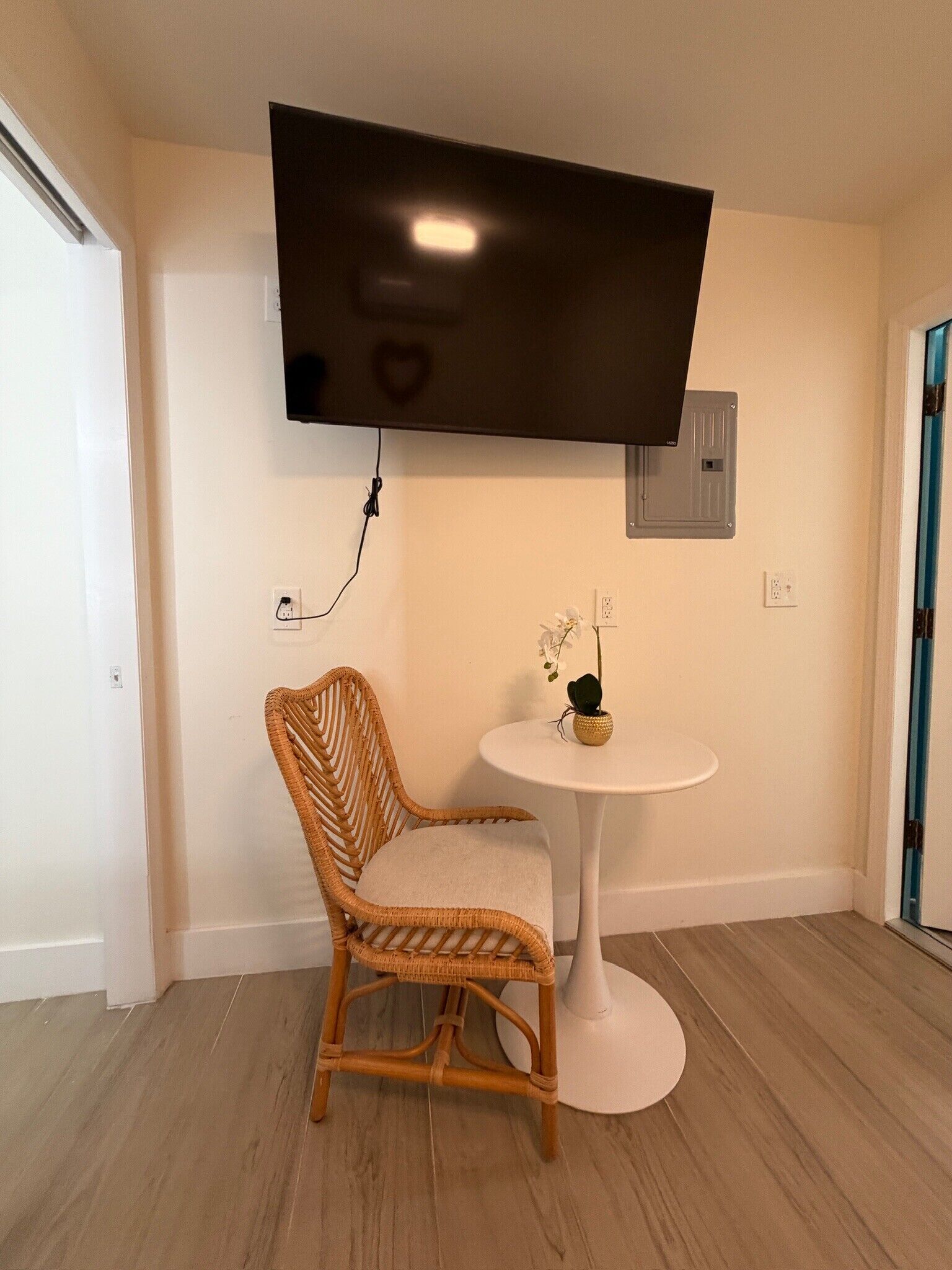 A room with a flat-screen TV mounted on the wall, a wicker chair, a small round table with a vase of flowers, and a door with a blue frame.