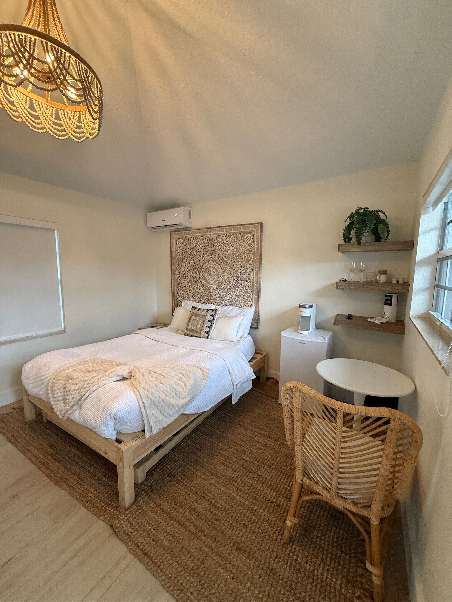 A bedroom with a bed, a round table, a wicker chair, a decorative wall panel, and a hanging light fixture.