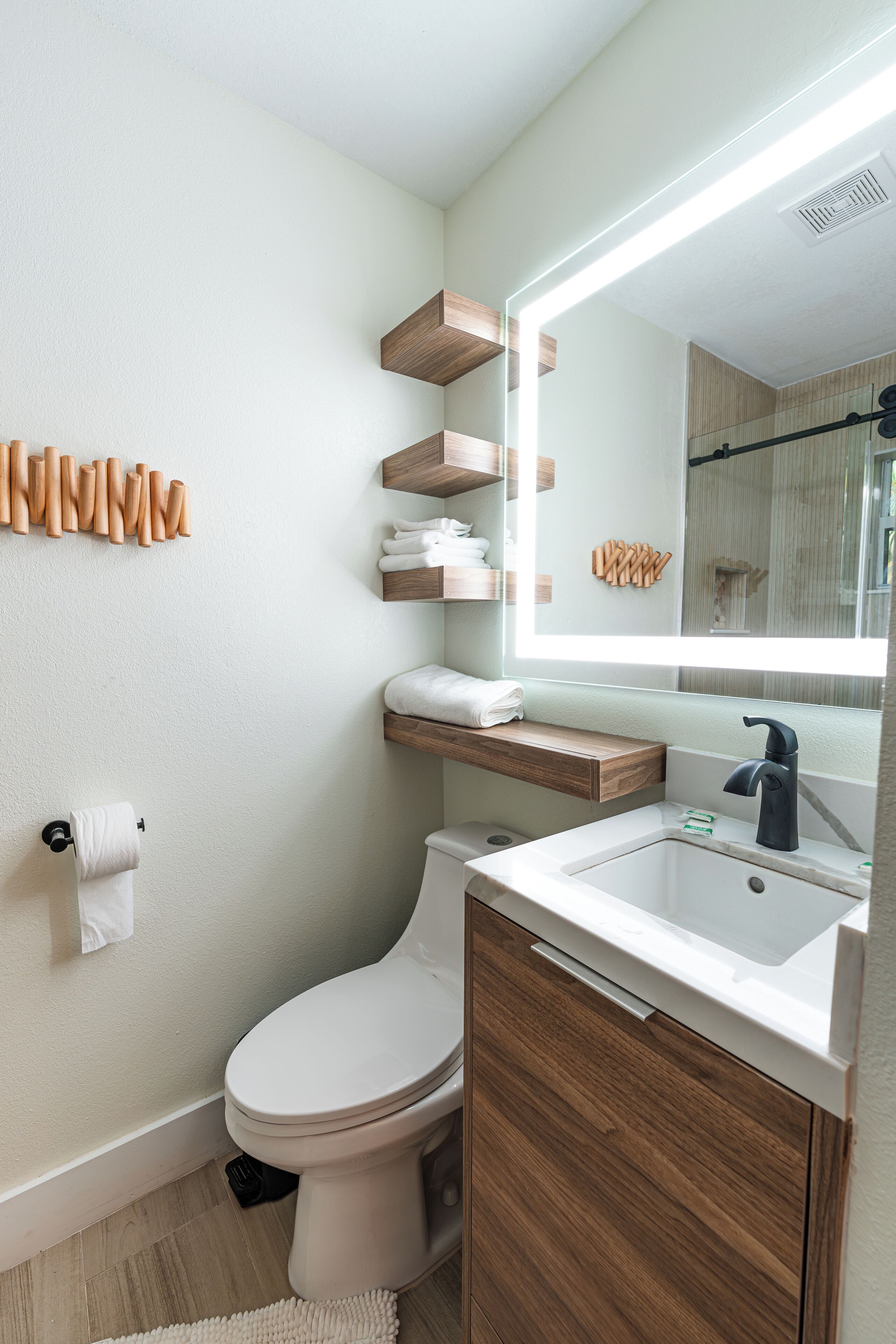 A modern bathroom with a toilet, sink, and mirror. There are wooden shelves above the sink and a towel rack on the wall.
