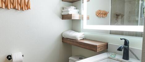 A modern bathroom with a toilet, sink, and mirror. There are wooden shelves above the sink and a towel rack on the wall.