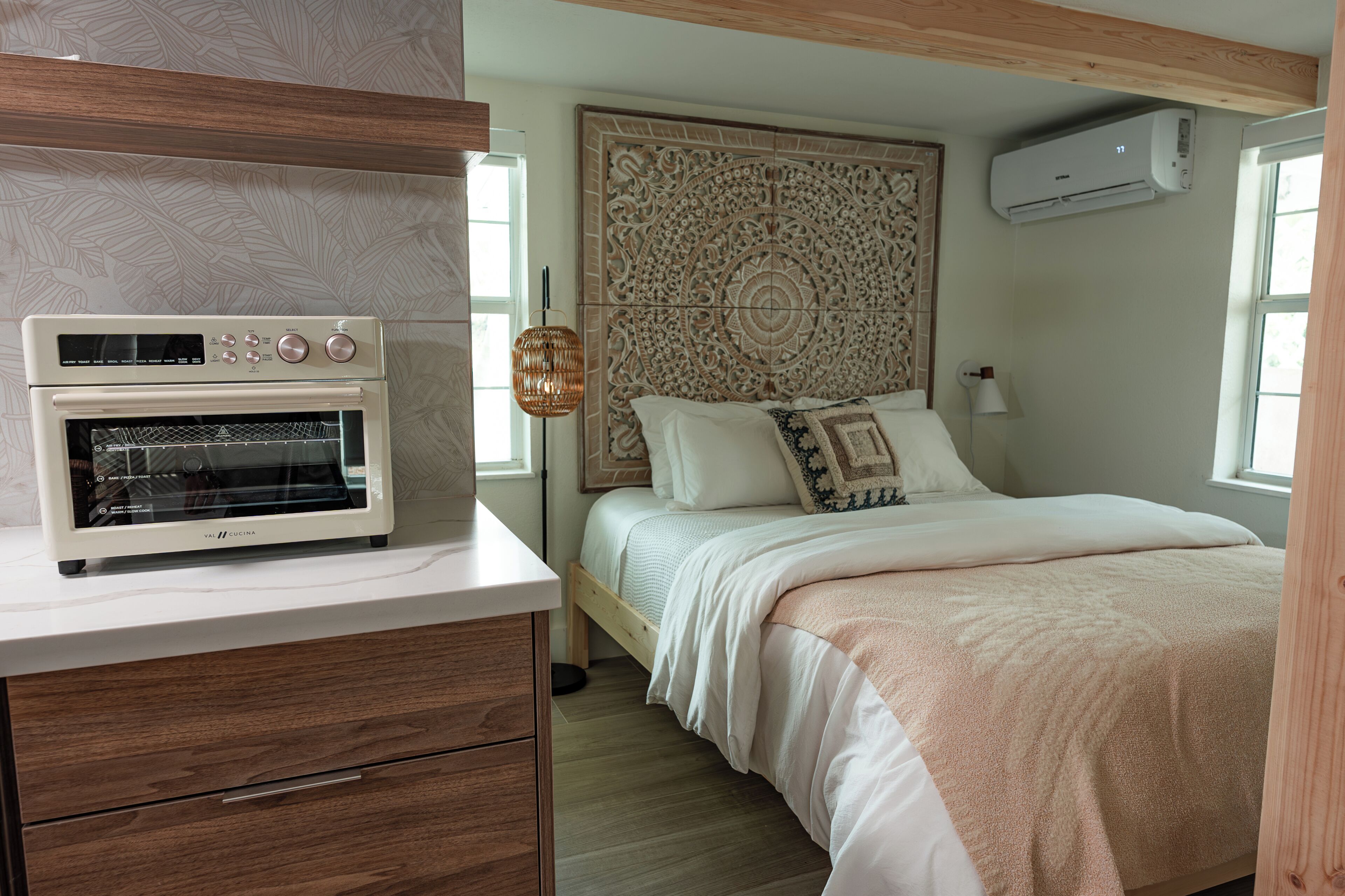 A bedroom with a bed, a wall-mounted air conditioner, a wooden headboard, a white bedside table with a built-in oven, and a window.