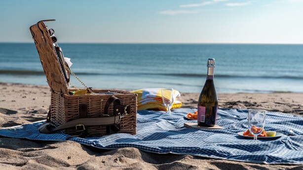 A picnic setup on a beach with a wicker basket, a bottle of wine, and a glass on a checkered blanket. - Seahorse Beach Bungalows (Fort Pierce)