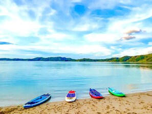 Una spiaggia con kayak disposti sulla sabbia, un lago tranquillo sullo sfondo e colline in lontananza.