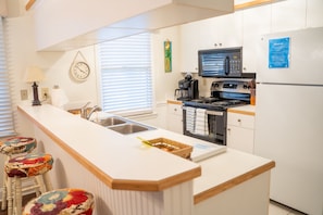 Una cocina moderna con muebles blancos, un lavamanos de acero inoxidable, un microondas, una estufa y un refrigerador. Hay taburetes coloridos en la isla de la cocina.