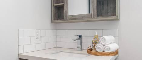 A bathroom with a white sink, a wooden-framed mirror, and a basket of rolled towels.
