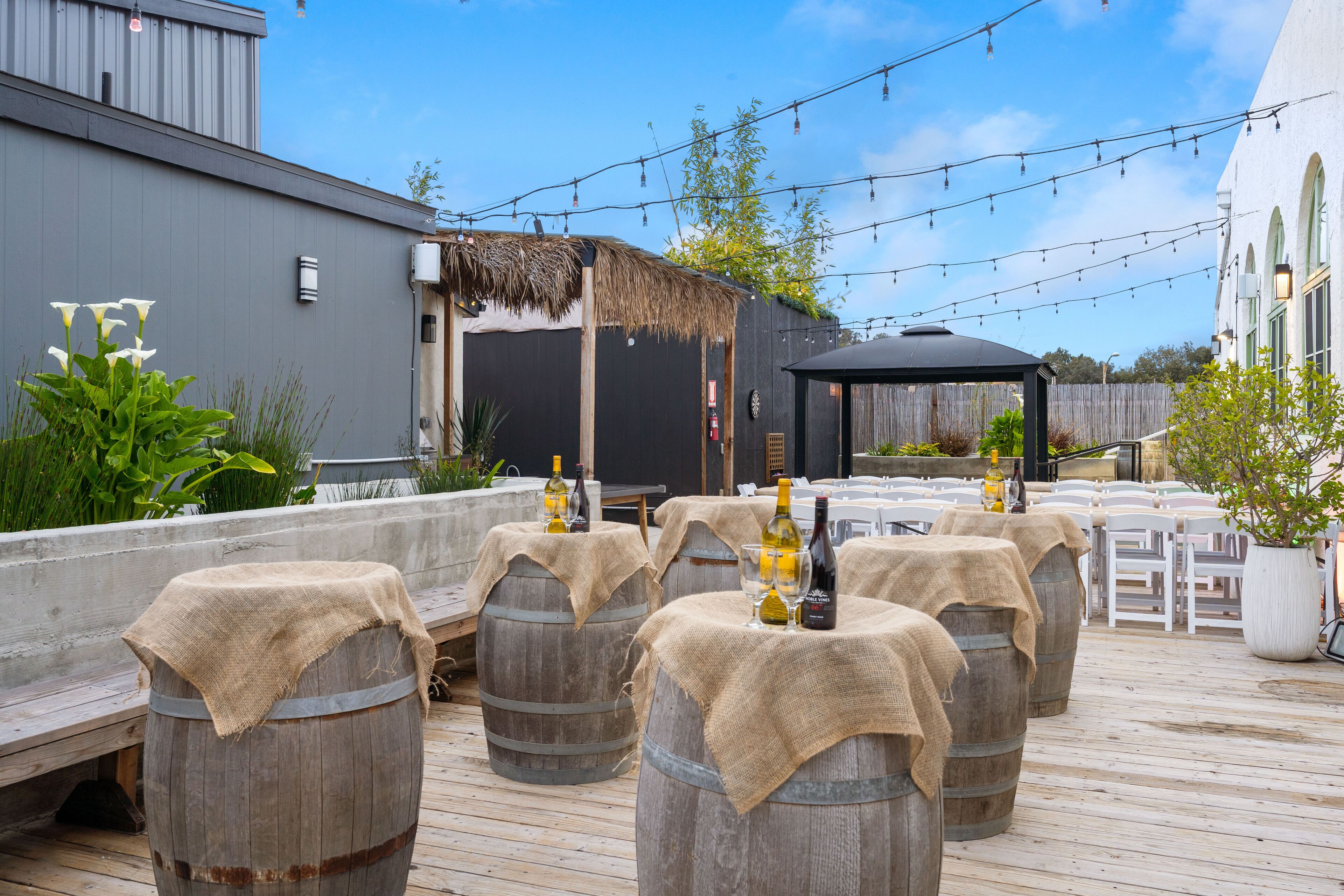 Una terraza de madera con barriles que sirven como mesas, luces colgantes y una estructura con techo de paja.