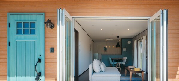 Ein modernes Interieur mit einer türkisblauen Tür, einer gemütlichen Sitzecke mit Sofa und Sesseln, einem kleinen Esstisch und einer Küchenzeile mit Kühlschrank und Pendelleuchten.