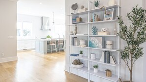 A modern kitchen with white cabinetry, a dining area with chairs, and a bookshelf filled with books and decorative items. - 5BR 4.5 BA Beach House close to beach on IOP with Private Pool! (Isle of Palms)