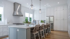 A modern kitchen with white cabinets, a stainless steel range hood, a central island with bar stools, and pendant lighting. - 5BR 4.5 BA Beach House close to beach on IOP with Private Pool! (Isle of Palms)