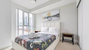 Un dormitorio moderno con una cama grande, vistas a la ciudad, un cuadro y una mesita de noche pequeña.