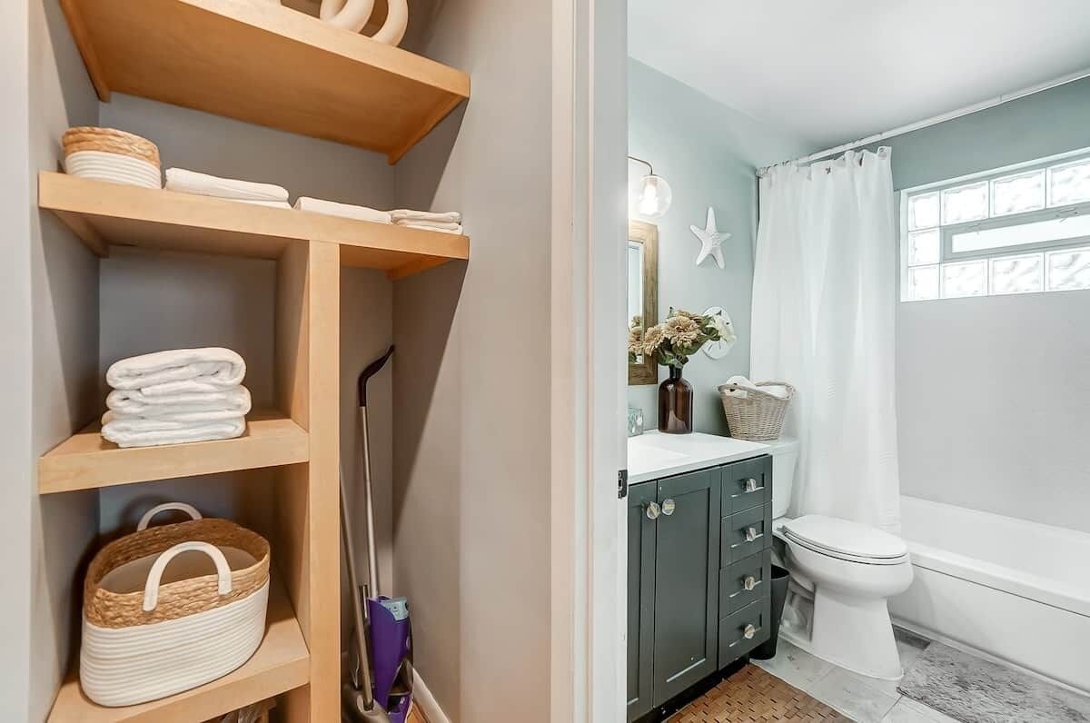A bathroom with a toilet, sink, and bathtub. There is a shelf with towels and a basket.