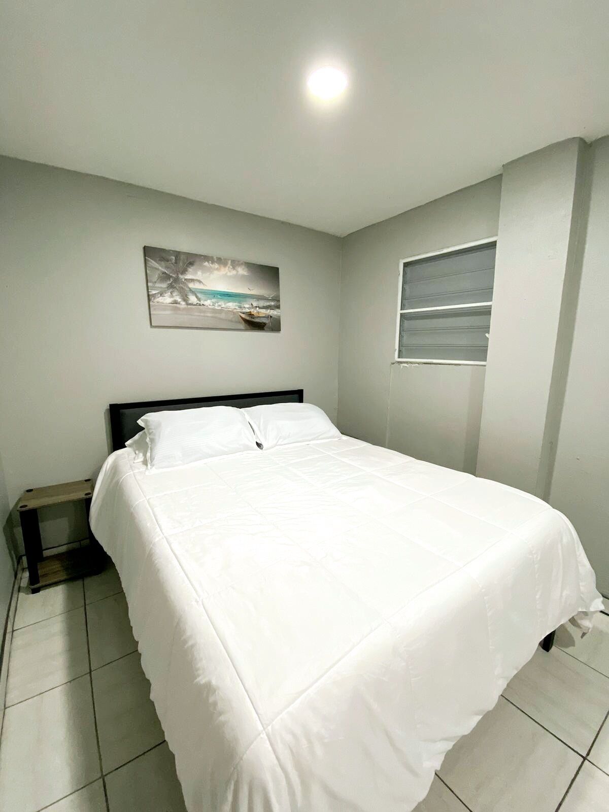 A neatly made bed in a room with a painting of a beach scene on the wall and a window with blinds.