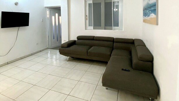 A living room with a sectional sofa, a flat-screen TV mounted on the wall, a window with blinds, and a door leading to another room. - Apartment Rio Nigua  (Salinas)