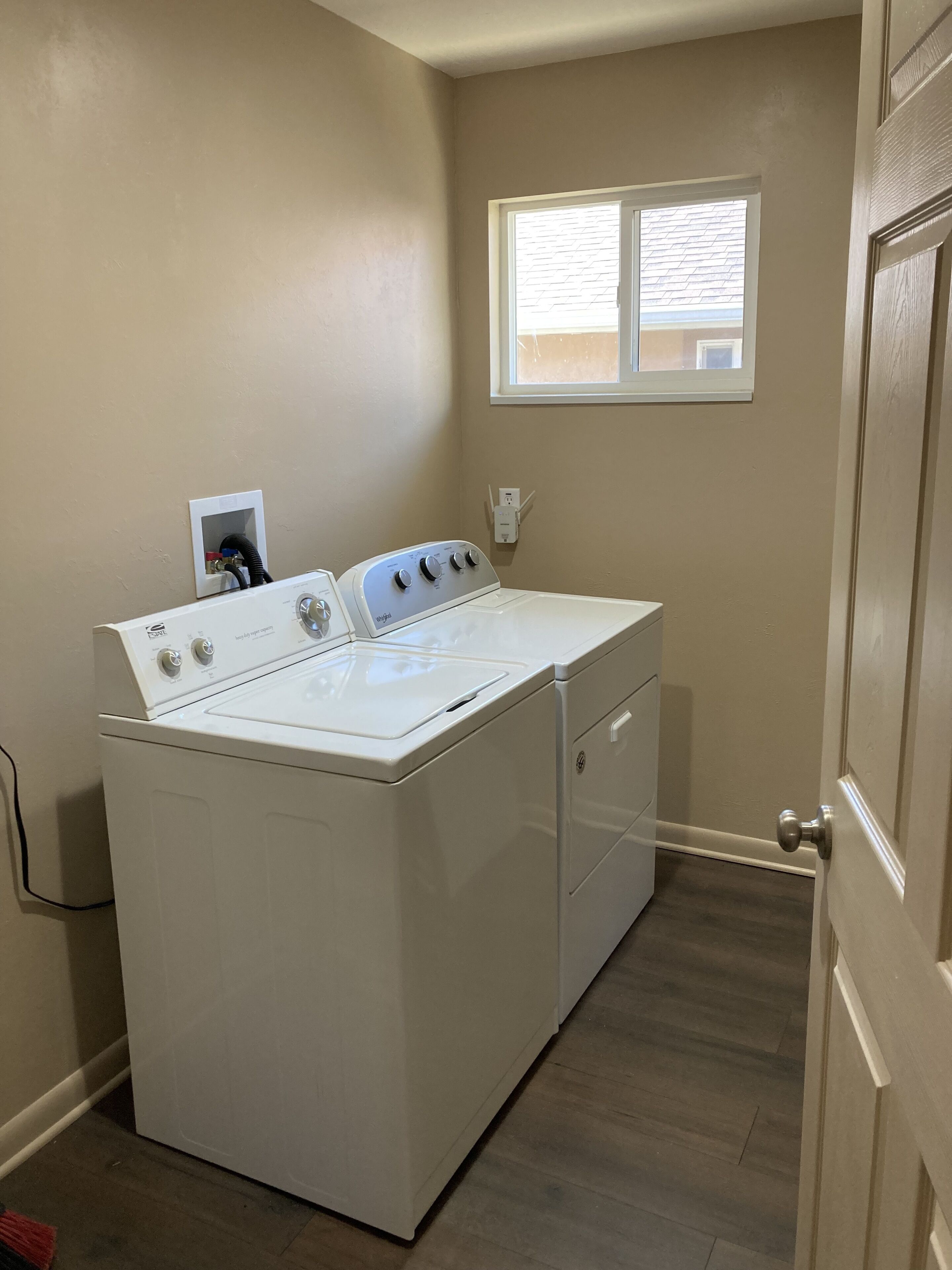 A laundry room with a front-loading washer and dryer, a window with blinds, and a door to the right.