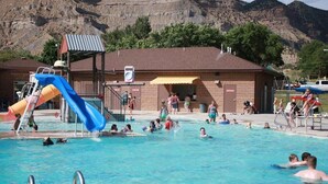 A swimming pool with a slide and a lifeguard tower, surrounded by a building with a brick facade and a mountain backdrop. - Cozy Home in Historic Helper Utah, near park, pool, Utah Aggies baseball field. (Helper)