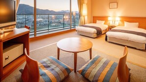 Ein Hotelzimmer mit großem Fenster und Blick auf die Stadt, einem Schreibtisch mit Fernseher, zwei Betten und einem kleinen runden Tisch.