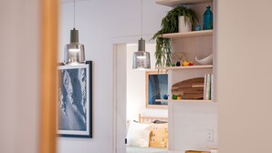 Ett modernt kök med vita bänkskivor, hängande taklampor, en inramad tavla på väggen och en trähylla med dekorativa föremål.